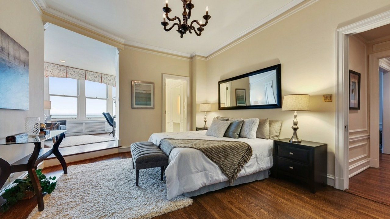 Beautiful bedroom in a Lake Shore Drive condo residence with king-size bed, nightstands, and bay window area with a desk. Art and mirror on walls, white bedding, throw blanket, and wall of throw pillows. White rug and contemporary style bench included.