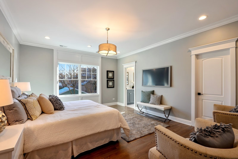 Hinsdale primary/master bedroom with king size bed, white bedding, wall of pillows, 2 nightstands, hanging drum light fixture, faux TV, white bench with pillows, pair of art prints, 2 matching accent chairs, and gray rug on dark maple floors.