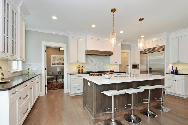 Downers Grove single family home kitchen with island, white cabinets, white marble countertops, 3 white contemporary barstools, fruits, canisters, bowls, and bottles on the countertops, and home office in background with accent chair, artwork, end table, and lamp.