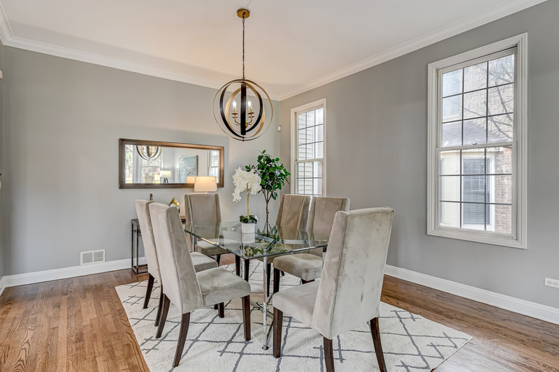 Hinsdale dining room staged with nail-headed accent chairs, large 6' glass dining table, rectangular mirror on the wall, faux tree in the corner, and large white dining rug with gray x's. The room has two windows and gray walls.