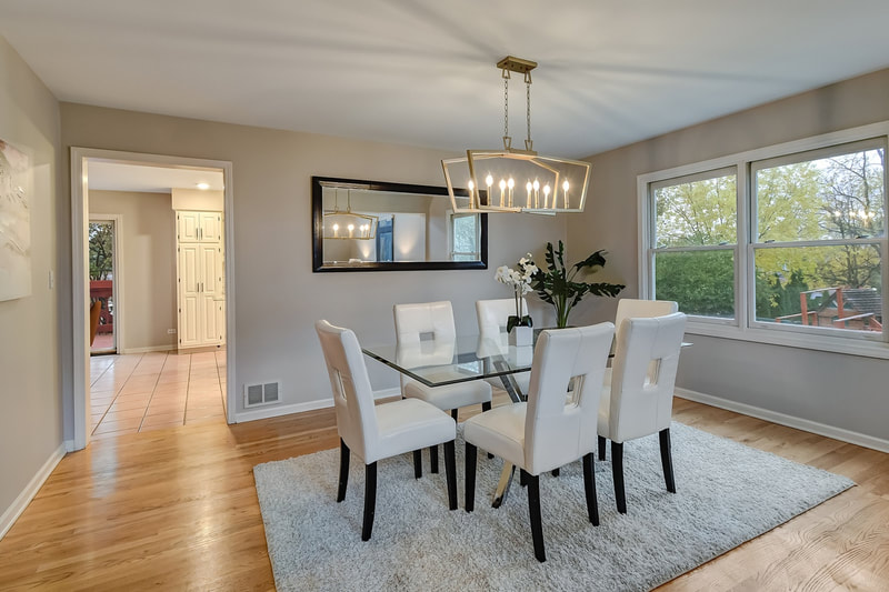 Oak Brook single family home's dining room staged with modern white chairs, glass table, contemporary light fixture, faux tree, and mirror on wall.