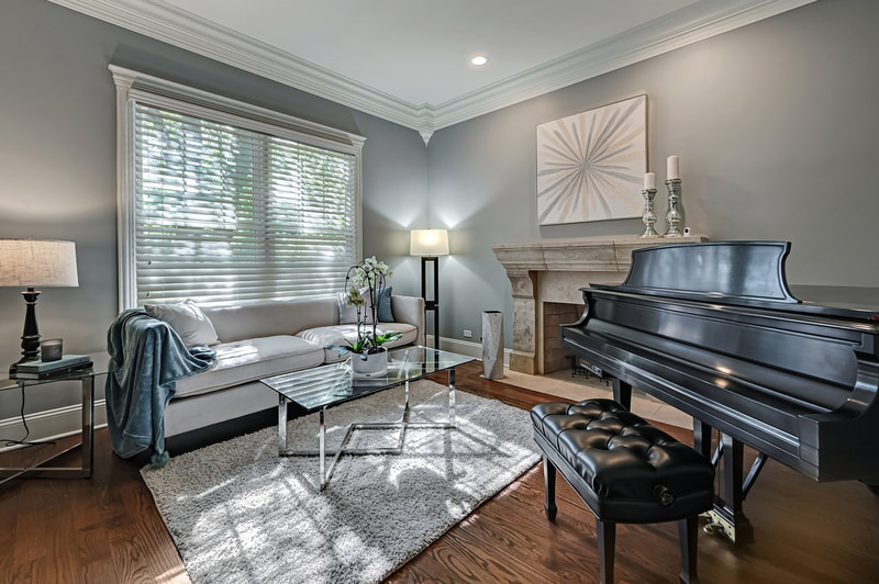 Sophisticated living space in an occupied home, staged to highlight its musical centerpiece, a grand black piano with a polished finish. The piano bench, upholstered in tufted black leather, stands ready for a performer. Natural light filters through Venetian blinds, casting a warm glow on the creamy sofa accented with a throw blanket. A textured white area rug anchors a glass coffee table, creating a harmonious blend of contemporary and classic elements. The traditional stone fireplace adds a timeless charm, complemented by a simple yet elegant piece of art and chic candlesticks atop the mantelpiece.