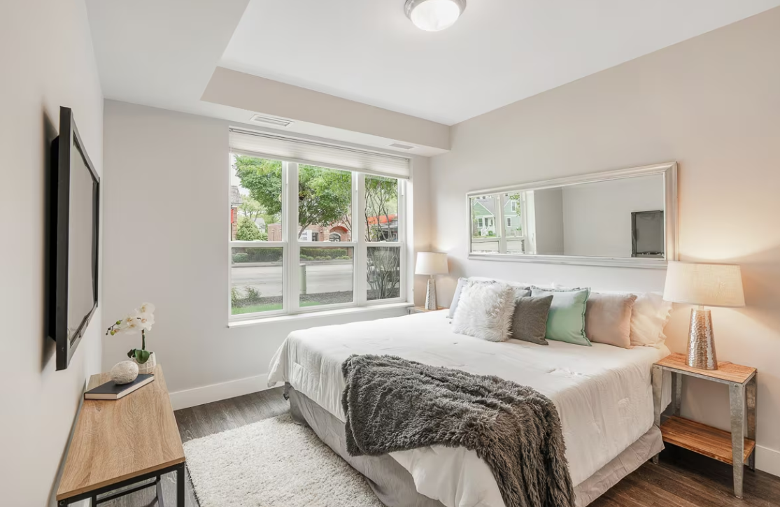 Elegantly staged modern bedroom with ample natural light. Features include a neatly made bed with white linens and a mix of gray and pastel green pillows, a cozy gray fur throw, two wooden bedside tables with decorative items such as a lamp and books, a large mirror across the bed, a wall-mounted flat-screen TV, and a soft white area rug. The room's large window offers a view of the neighborhood, inviting a serene atmosphere.
