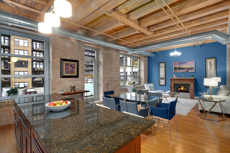Timber Loft in Chicago with contemporary blue dining chairs and glass table. Living room includes combined dining space, blue accent wall with TV over fireplace, and pair of art prints on either side of the fireplace. Kitchen island included in the photo.