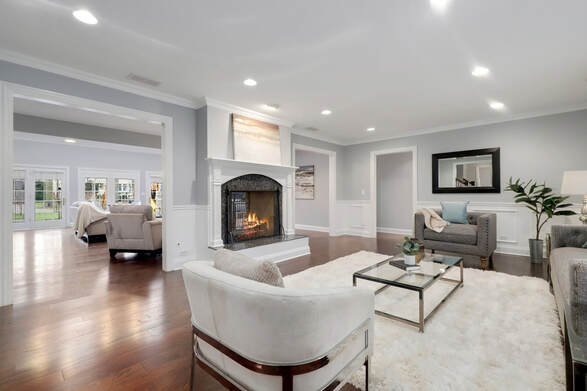 PictureElegantly staged living room with hardwood floors, white trim, and gray walls. The room features a white fireplace with a burning fire, flanked by two framed artworks. Two white armchairs and a gray sofa are arranged around a glass coffee table over a plush white area rug. The space is well-lit with recessed lighting, and the open layout leads to an adjoining room with a glimpse of outdoor patio doors.