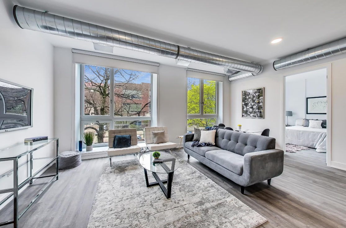 Contemporary urban living room in a model unit featuring exposed silver ductwork on the ceiling that adds an industrial vibe. Two large windows let in ample natural light, highlighting the room's grey-toned hardwood floors and the soft textures of the area rug. A tufted grey sofa, paired with a modern beige accent chair, provides comfortable seating. The glass coffee table with a geometric black base sits centrally, while tasteful artworks and sleek shelving units adorn the walls. The open floor plan allows a glimpse into the adjoining bedroom, showcasing a cohesive design aesthetic.